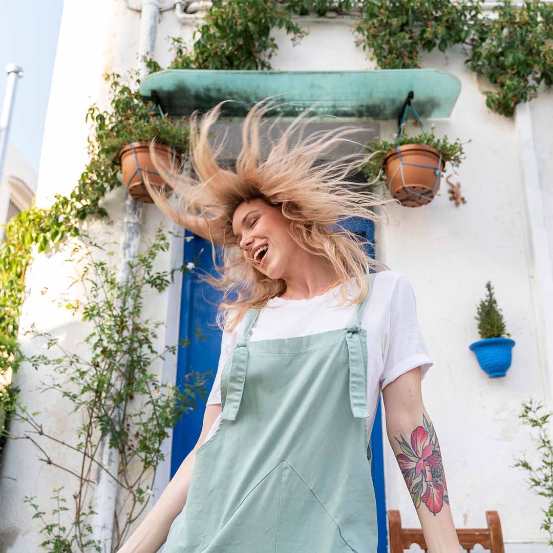 Junge Frau mit wehendem Haar in mintgrüner Latzschürze, weißem T‑Shirt und großem Blumen‑Tattoo vor blauem Türrahmen