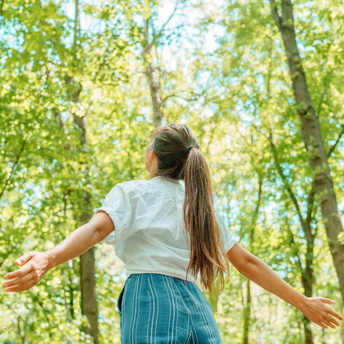Eine Frau mit langen braunen Haaren, weißem Hemd und blauer Hose steht mit ausgebreiteten Armen im sonnigen, grünen Wald.