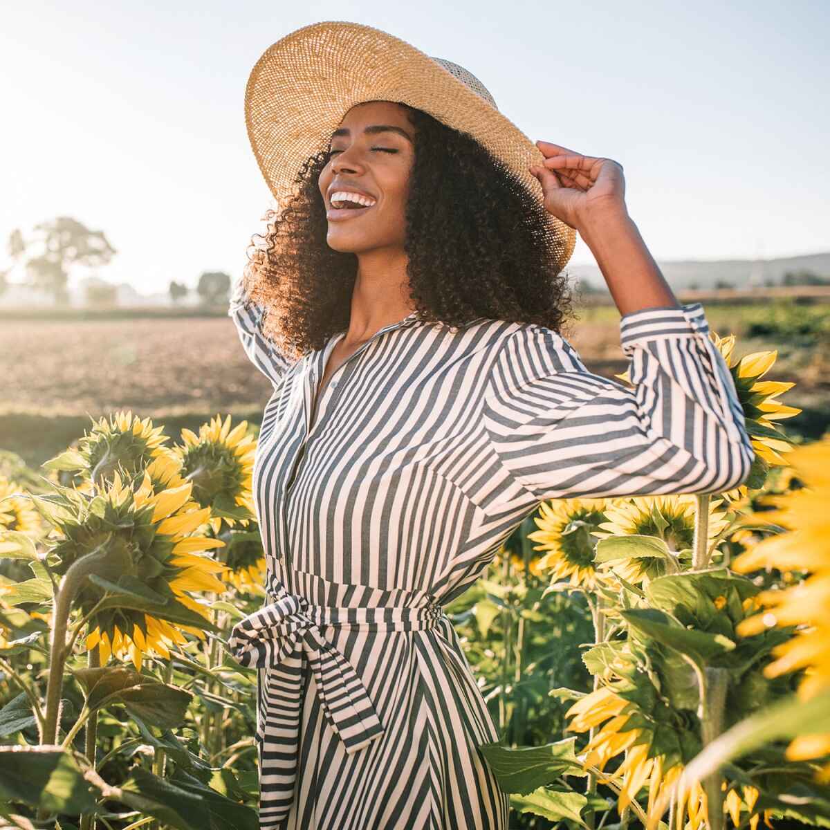 Eine lachende Frau mit lockigen Haaren trägt ein gestreiftes Kleid und einen Strohhut, steht inmitten blühender Sonnenblumen auf einem Feld bei klarem, sonnigem Wetter. Im Hintergrund sieht man unscharfe Felder und einen blauen Himmel.