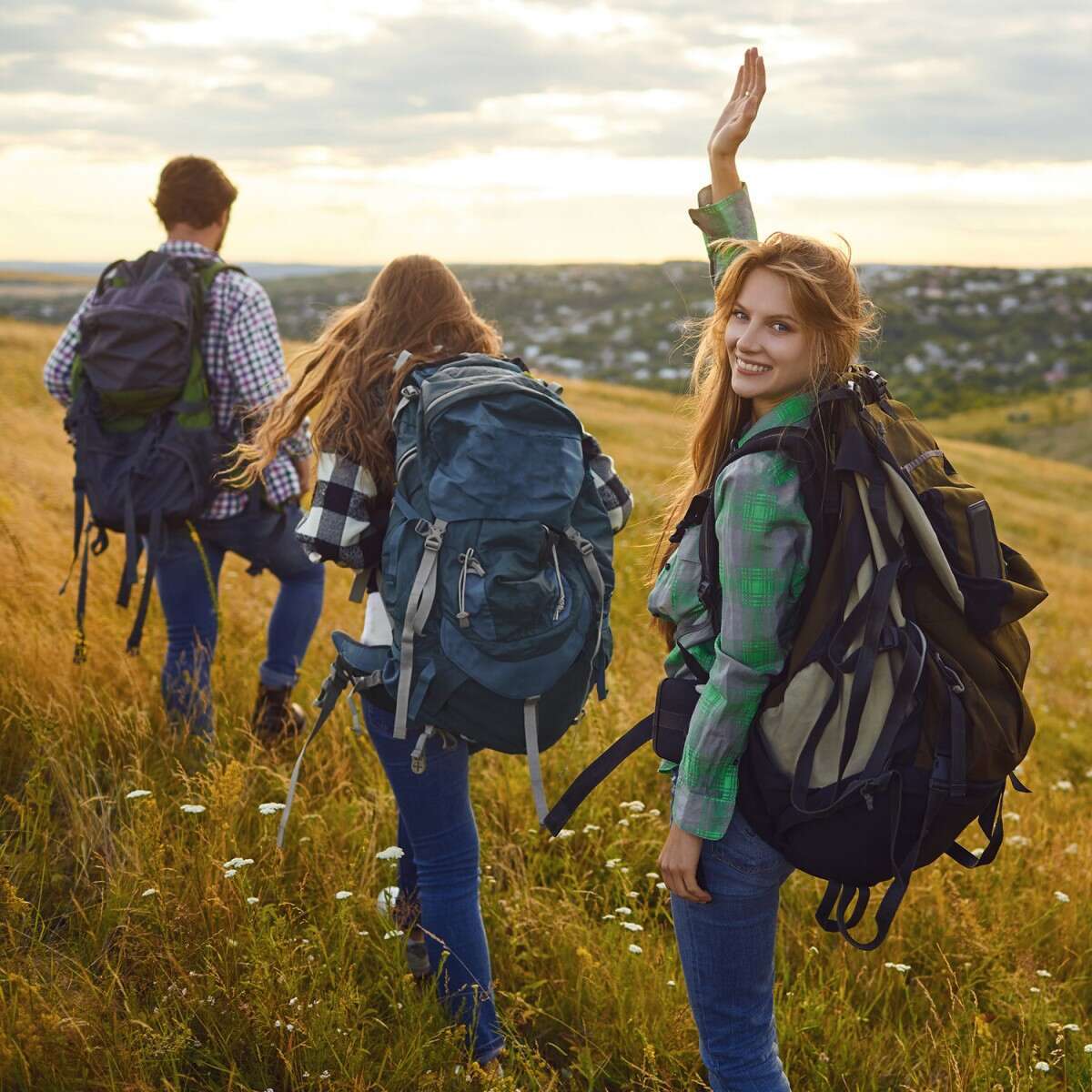 Drei junge Menschen mit großen Wanderrucksäcken wandern durch eine Wiese, im Hintergrund hügelige Landschaft und blauer Himmel.