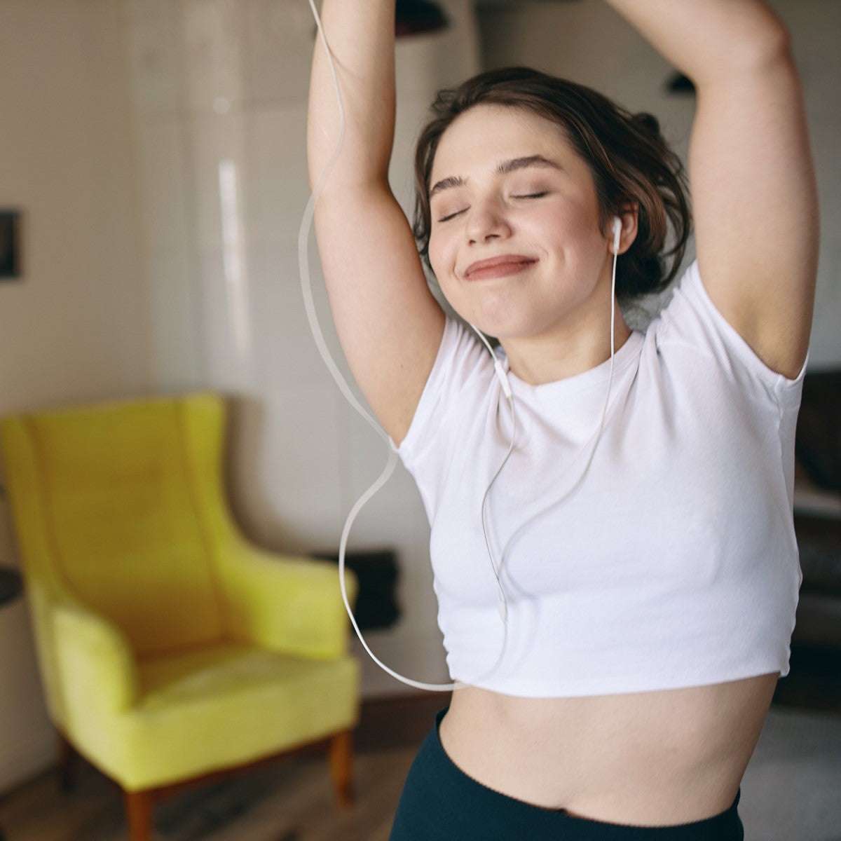 Eine junge Frau mit weißem T-Shirt und Kopfhörern hebt die Arme, lächelt entspannt, im Hintergrund ein gelber Sessel.