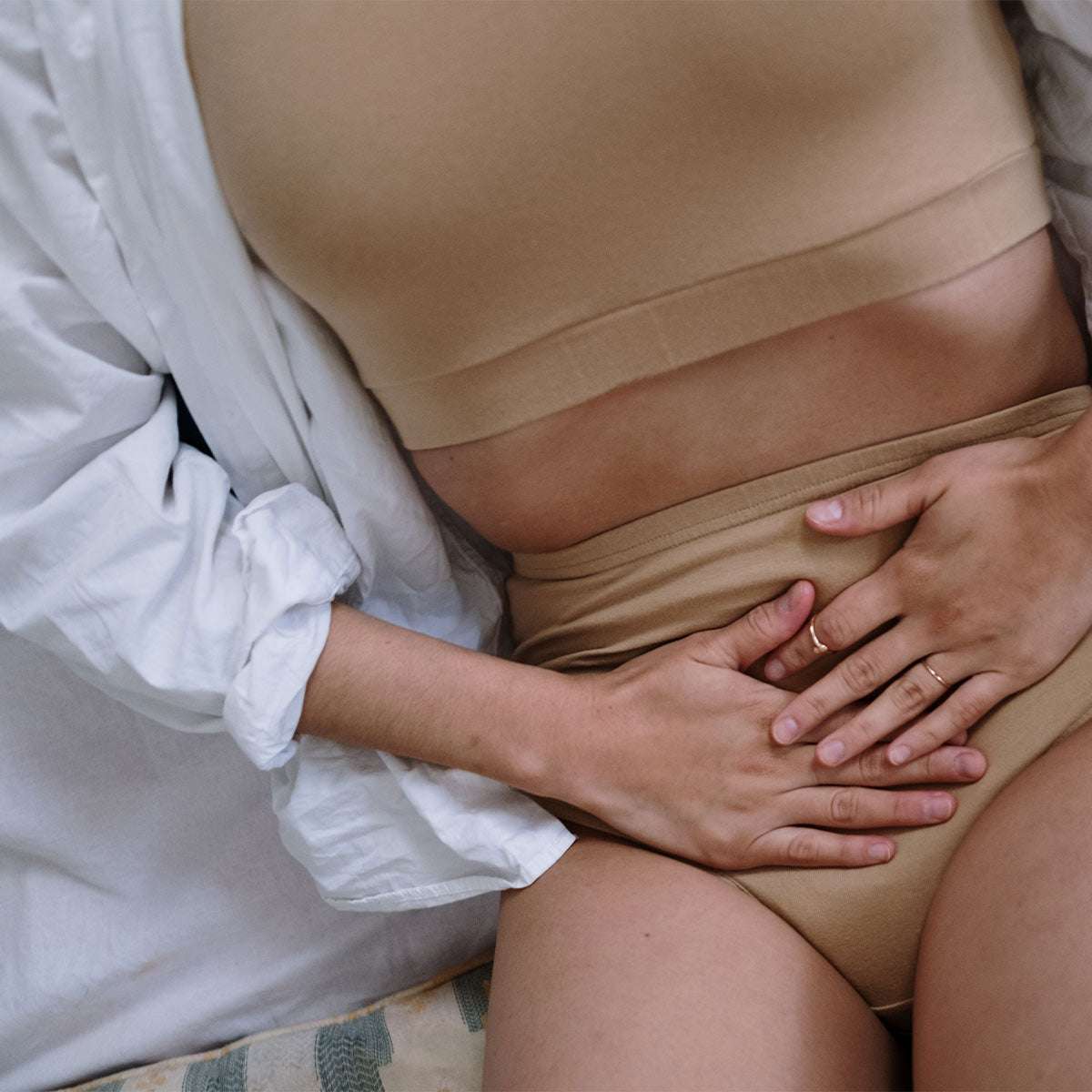 Eine Person trägt hautfarbene Unterwäsche und ein passendes Oberteil, liegt halb auf einer hellen Decke und hält beide Hände auf den Unterbauch. Eine weiße offene Bluse bedeckt einen Arm, die Person trägt zwei schmale Ringe an den Fingern. Die Szene wirkt entspannt, möglicherweise in einem Schlafzimmer.