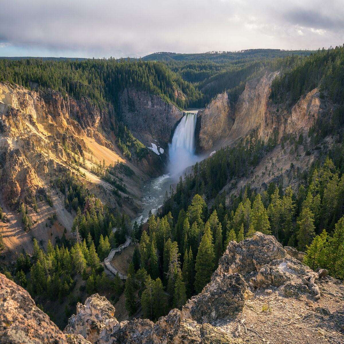 Im Vordergrund befinden sich Felsen, dahinter dichter grüner Nadelwald. Zentral fließt ein breiter Wasserfall in eine tiefe Schlucht. Die Felswände sind links gelblich, rechts grau-orange. Im Hintergrund erstrecken sich ausgedehnte Wälder unter bewölktem Himmel. Ein Wanderweg ist unten im Wald sichtbar.